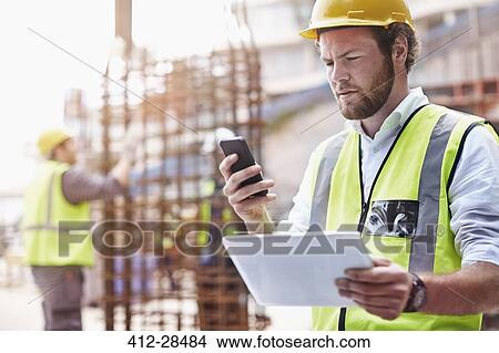 Picture - Construction worker with digital tablet texting with cell phone at construction site. Fotosearch