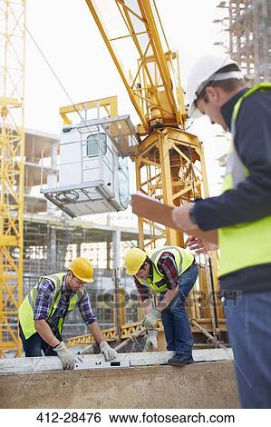 Stock Photograph - Construction workers using level tool below crane at construction site. Fotosearch