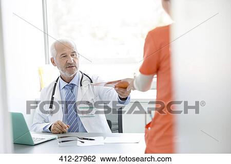 Stock Image - Doctor at desk giving medical record to nurse in doctor’s office. Fotosearch