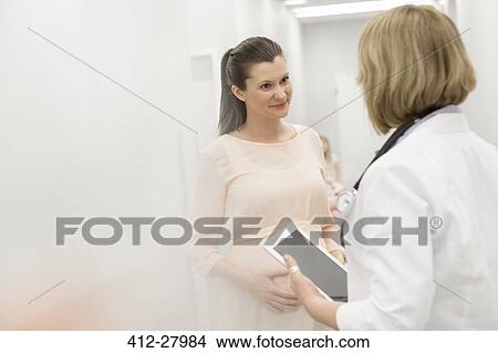 Doctor with digital tablet talking to pregnant patient in clinic corridor View Large Photo Image Picture - Doctor with digital tablet talking to pregnant patient in clinic corridor. Fotosearch
