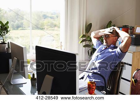 Man leaning back working at computer in sunny home office View Large Photo Image Stock Photo - Man leaning back working at computer in sunny home office. Fotosearch