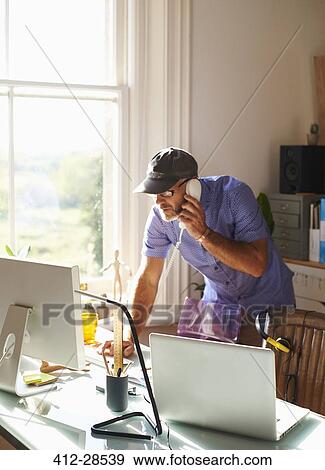 Stock Photo - Man talking on telephone and using computer at desk in sunny home office. Fotosearch