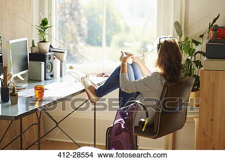 Pensive woman looking through window with feet up on desk in sunny home office View Large Photo Image Picture - Pensive woman looking through window with feet up on desk in sunny home office. Fotosearch