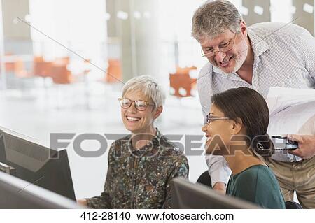Stock Image - Smiling students talking at computer in adult education classroom. Fotosearch