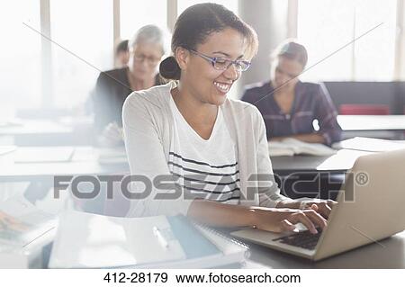 Stock Photo - Smiling woman typing at laptop in adult education classroom. Fotosearch