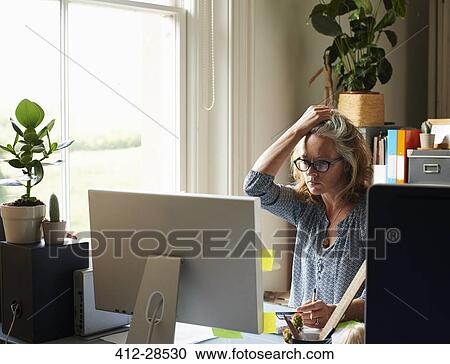 Stock Image - Stressed woman with hand in hair holding credit card at computer in home office. Fotosearch