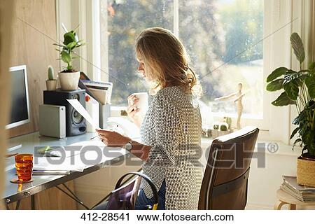 Stock Image - Woman drinking coffee and reading paperwork at desk in sunny home office. Fotosearch