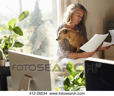 Picture - Woman with dog reviewing paperwork in sunny home office. Fotosearch
