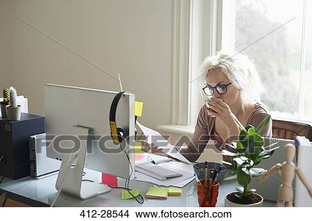 Picture - Young woman drinking tea and reading paperwork at desk in home office. Fotosearch