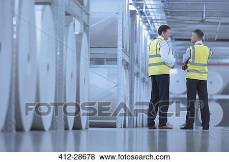 Stock Photo - Businessmen in reflective clothing talking near large paper spools in printing plant. Fotosearch