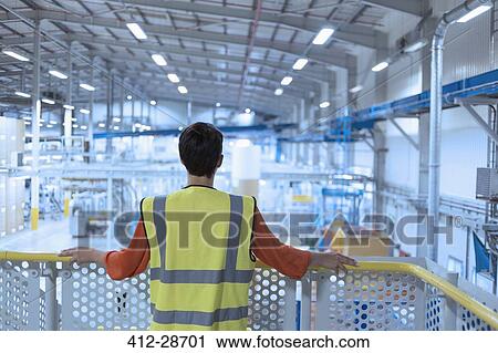 Stock Image - Worker in reflective clothing on platform looking out over factory. Fotosearch