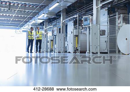 Stock Photo - Workers walking along machinery in factory. Fotosearch
