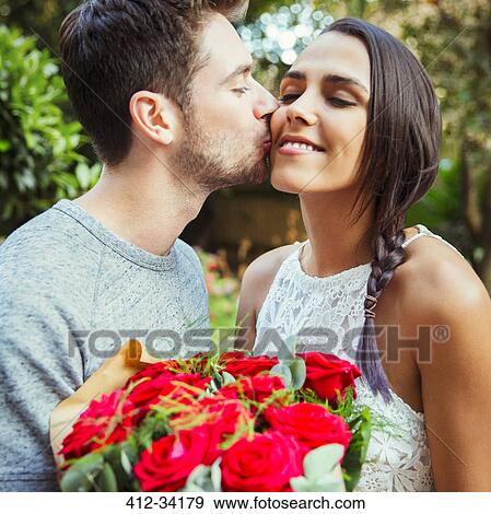 Stock Photo - Affectionate man giving red rose bouquet to girlfriend, kissing her on cheek. Fotosearch