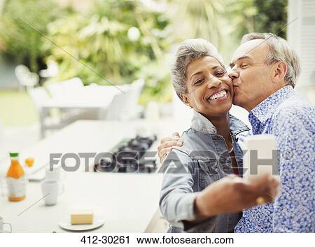 Stock Image - Affectionate mature couple kissing taking selfie in kitchen. Fotosearch