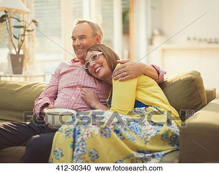 Stock Image - Affectionate mature couple watching TV and eating popcorn on living room sofa. Fotosearch