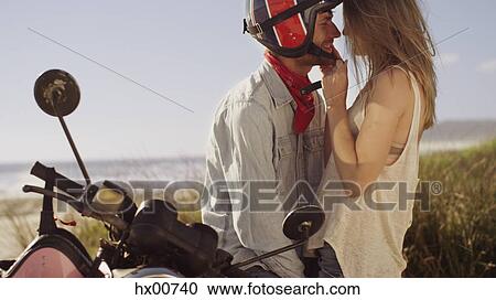 Stock Image - Affectionate young couple at motorcycle with beach in background. Fotosearch