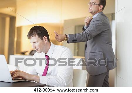 Picture - Angry businessman aiming rubber band at unsuspecting businessman working at laptop. Fotosearch