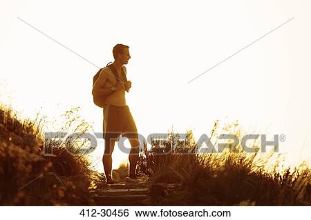 Stock Photograph - Bare chested male hiker with backpack on trail at sunset. Fotosearch
