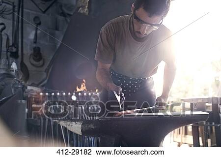 Stock Image - Blacksmith forging hot iron at anvil in forge. Fotosearch