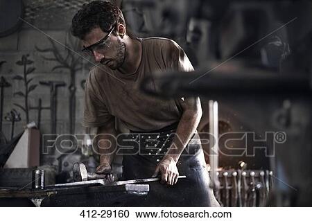 Stock Image - Blacksmith hammering iron at anvil in forge. Fotosearch