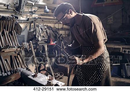 Stock Image - Blacksmith heating metal with blowtorch in forge. Fotosearch