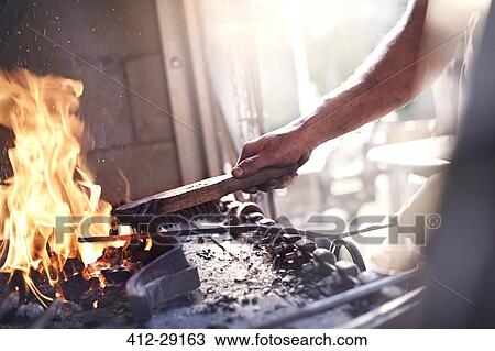 Stock Image - Blacksmith heating tool at fire in forge. Fotosearch