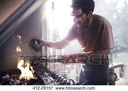 Stock Photo - Blacksmith pouring liquid over tool at fire in forge. Fotosearch