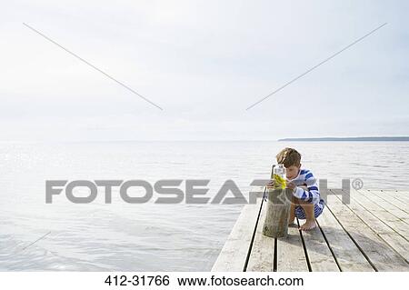Stock Photograph - Boy examining seaweed in jar on sunny lakeside dock. Fotosearch
