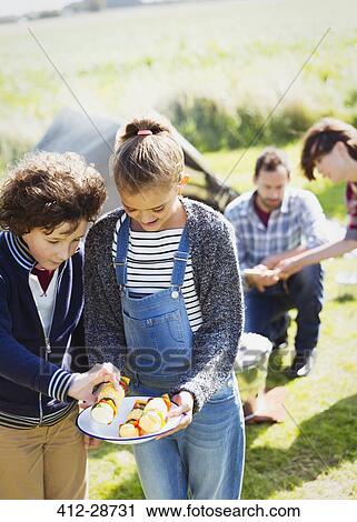 Stock Image - Brother and sister looking at vegetable skewers at sunny campsite. Fotosearch