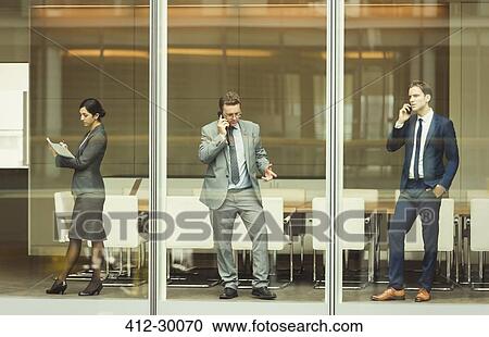 Stock Image - Business people on cell phones and taking notes at conference room window. Fotosearch