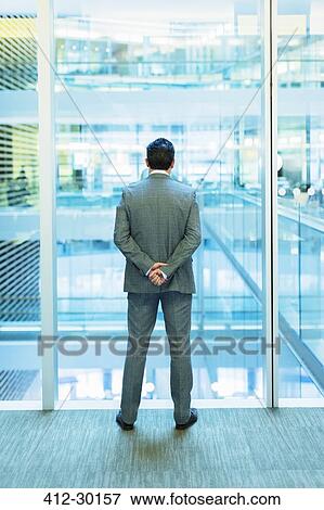 Stock Photo - Businessman standing at atrium window with hands behind back. Fotosearch