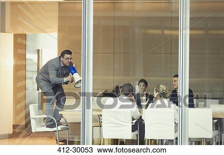 Stock Image - Businessman with megaphone on top of chair in conference room meeting. Fotosearch
