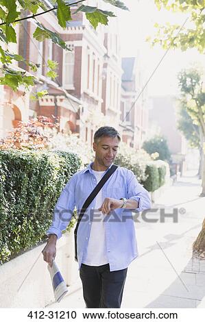 Stock Image - Businessman with newspaper walking on sidewalk and checking wristwatch. Fotosearch