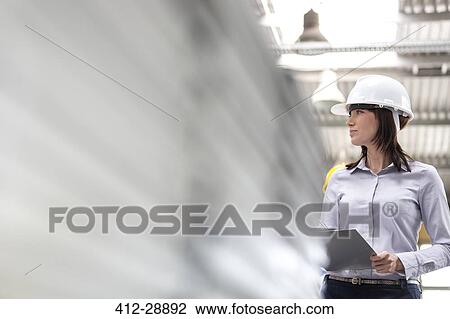 Stock Image - Businesswoman in hard-hat and clipboard working in factory. Fotosearch