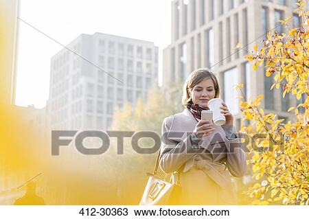 Stock Image - Businesswoman texting and drinking coffee in city. Fotosearch