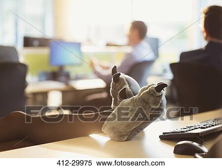 Stock Photography - Businesswoman wearing wolf paw slippers with feet up on office desk. Fotosearch