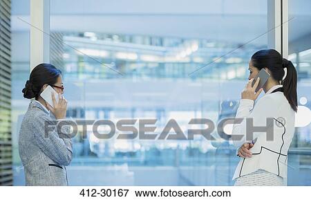 Stock Photo - Businesswomen talking on cell phones at office window. Fotosearch