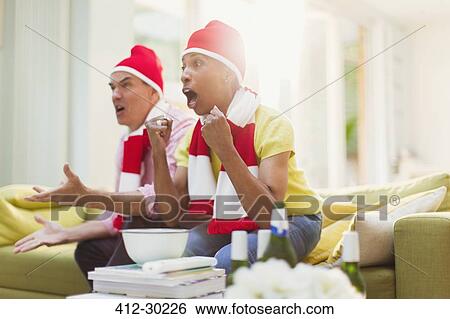 Stock Photograph - Cheering mature couple wearing matching hats and watching TV sports event in living room. Fotosearch