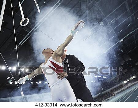 Coach lifting male gymnast below gymnastics rings in arena View Large Photo Image Picture - Coach lifting male gymnast below gymnastics rings in arena. Fotosearch