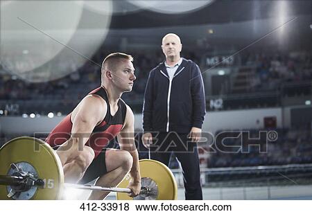Coach watching focused male weightlifter lifting barbell in arena View Large Photo Image Stock Photo - Coach watching focused male weightlifter lifting barbell in arena. Fotosearch