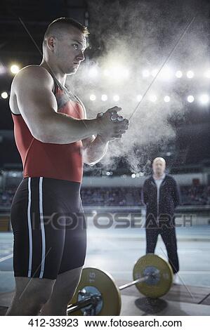 Stock Image - Coach watching male weightlifter applying chalk powder to hands at barbell. Fotosearch