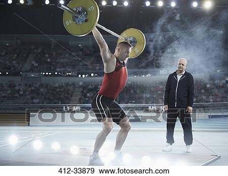 Coach watching male weightlifter squatting barbell overhead in arena View Large Photo Image Stock Photo - Coach watching male weightlifter squatting barbell overhead in arena. Fotosearch