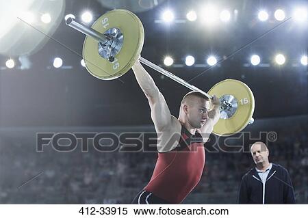Stock Photography - Coach watching male weightlifter squatting barbell overhead in arena. Fotosearch