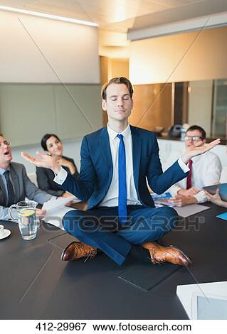 Stock Photo - Colleagues watching zen-like businessman meditating in lotus position on conference table. Fotosearch