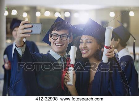 Stock Photo - College graduates in cap and gown holding diplomas posing for selfie. Fotosearch