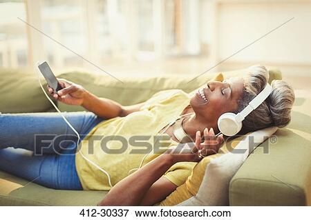 Stock Photo - Comfortable mature woman listening to music with headphones and mp3 player on living room sofa. Fotosearch