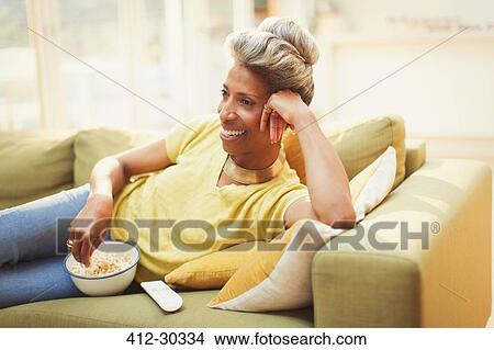 Comfortable mature woman watching TV and eating popcorn on living room sofa View Large Photo Image Picture - Comfortable mature woman watching TV and eating popcorn on living room sofa. Fotosearch