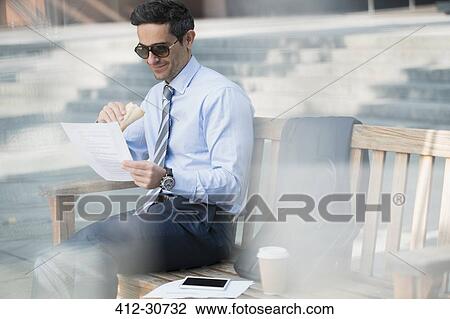 Stock Image - Corporate businessman eating lunch and working on bench. Fotosearch