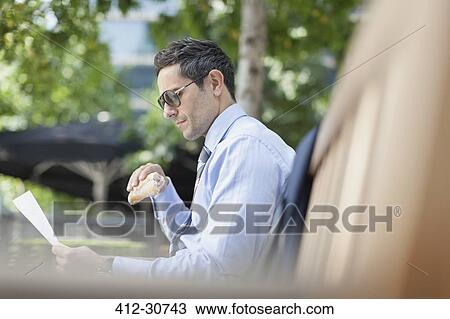 Corporate businessman eating lunch and reading paperwork on park bench View Large Photo Image Stock Image - Corporate businessman eating lunch and reading paperwork on park bench. Fotosearch