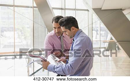 Corporate businessmen reviewing paperwork in modern office lobby View Large Photo Image Stock Photo - Corporate businessmen reviewing paperwork in modern office lobby. Fotosearch